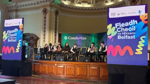 Seven kids are sitting on a brown wooden stage. Each side of them are two large posters promoting “Fleadh Cheoil ne hEireann Belfast.” Th kids on stage have an assortment of instruments in their hands. Behind them the walls are cream, they sit below a dome in the in a grand hall. Behind them is a green screen with the words Comhaltas.  
