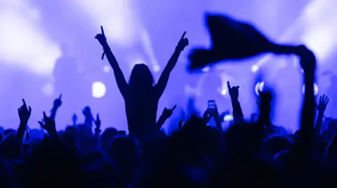 A generic scene of revellers reacting to live music. One person is shown on the shoulders of another rising out of a crowd of people with the hands in the air while a band plays in the background.
