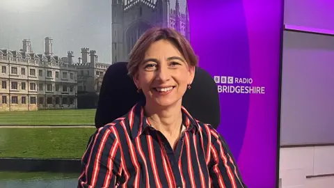 Sue Dougan/BBC Elizabeth McWilliams stares at the camera. She wears a black and red stripe dress. She sits on a chair in front of a sign that says BBC Radio Cambridgeshire and a picture of King's College chapel