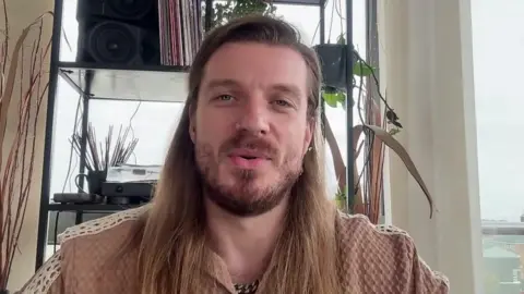 BBC News Dom Topix, who has long blond hair, sits in the living room talking to the BBC.