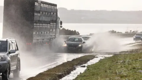 PA Media A black car drives through flood water on the A9, spraying it in all directions. A lorry is driving in the opposite direction in the next lane, while another car is just behind the first car.  