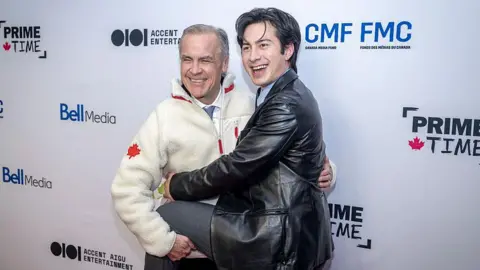 Getty Images Prime Minister Mark Carney, wearing a beige fleece sweater with a red Canadian maple leaf on the arm, laughs as he hugs Heated Rivalry actor Hudson Williams, wearing a leather jacket.