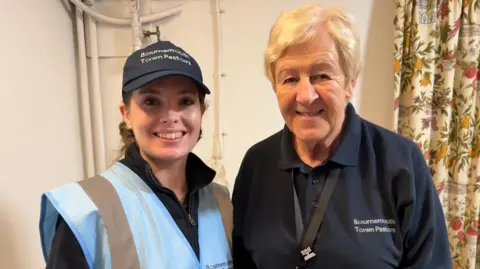 Two women wearing blue and smiling. One is wearing a cap that reads "Bournemouth Town Pastors."