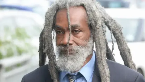 Judah Adunbi is standing, looking away from the camera wearing a navy blue suit, a light blue shirt and a dark blue tie. He has a grey beard and grey dreadlocks. Behind him are cars.