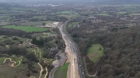 A drone image of an A-road, stretches of which are covered in construction sand as new parts are built, with vehicles travelling up and down it. The road is in a valley between two hills in the Cotswolds, with a biking track snaking up one of the hills. Green fields and houses are scattered in the distance.