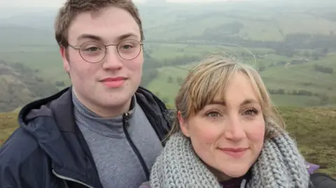 Woman with blonde hair and grey scarf with son, teenage boy with short brown hair and glasses wearing black coat, pictured infront of a hillside landscape 