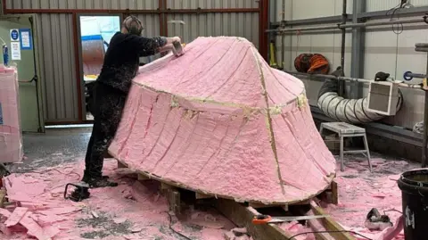 Coastal Forces Heritage Trust A man works on the bottom of the sculpture in the boathouse. It is currently pink.
