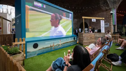 David Cleveland A photograph of a big screen being used outside to show a tennis match, with people watching in deckchairs. There is a bar to one side of the screen and people are wearing sunglasses and eating ice cream.