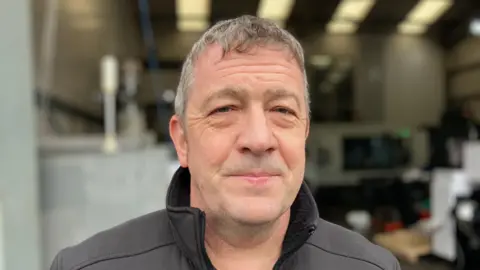 BBC Man with this grey curly hair is looking directly at the camera and wears a black high collar jacket