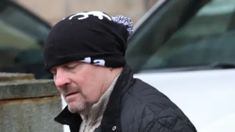 Tim Bugler Kevin Dolan at court, wearing a black jacket and a black hat covering part of his head. He has a moustache and trim beard.