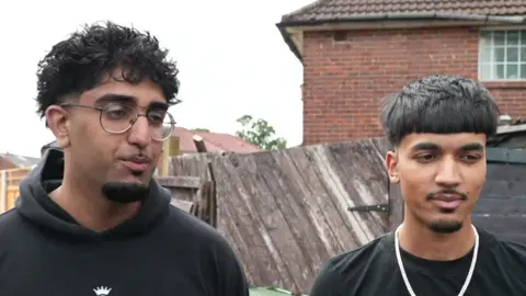 Two young men with black hair and beards stand in front of a fence.