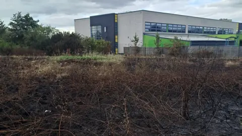 MFRS A view across a charred and blackened field. The scorch marks reach up to a fence, separating the field from a large, grey flat-roofed building. 