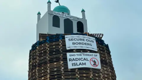 BBC An image of the top of the bonfire, consisting of wood pallets, with a structure resembling a mosque on top. Two placards are below. One reads "secure our borders" while another reads "end the threat of radical Islam".