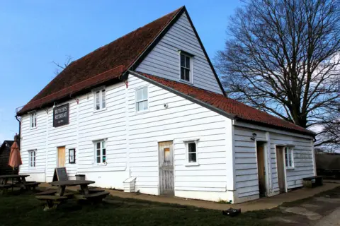 Historic England A white wooden slat building which has a red tiled roof. It has benches and tables in front of it and a tree behind it