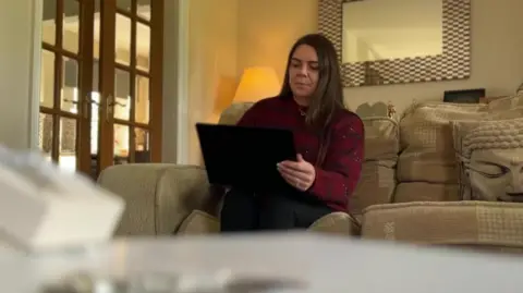 BBC/NATHAN TURVEY A picture of Madeline Roberts who has dark brown long hair and is sitting on her sofa looking at her laptop, there is a mirror, lamp and doors behind her.