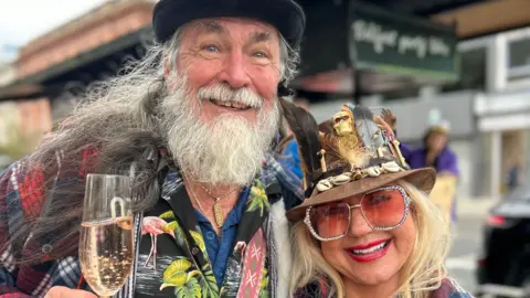 Melody and John Hennessee in Belfast smiling and holding glasses of prosecco. They are wearing cowboy hats and fun glasses.