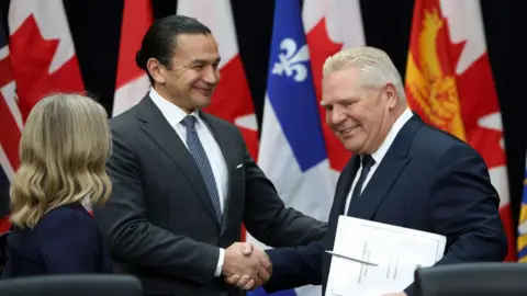 Bloomberg via Getty Images Manitoba Premier Wab Kinew and Ontario Premier Doug Ford, both in dark suit, smile as they shake hands during a meeting of premiers. Behind them is a number of flags representing different provinces. A blond woman stands with her back to the camera on the left. 