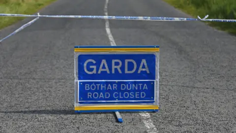 A blue bilingual road sign which reads:" Garda - bothar dunta - road closed."
The sign in sited in the middle of an empty road, under a cordon made of blue and white tape
