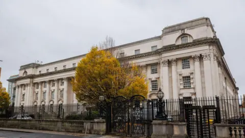 PA Media The Belfast High Court is a stone coloured building in Belfast with black metal fences and gates around it. There is a green tree in front of the building.