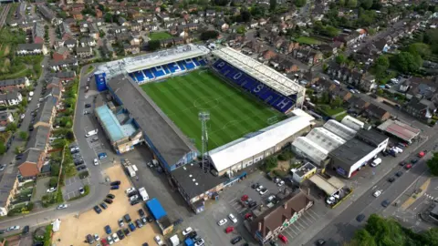 An aerial view of Weston Homes Stadium . The pitch is surrounded by blue seats with white seats which spell out '1939 Posh'. Around the stadium there are two car parks as well as several streets and rows of houses. 