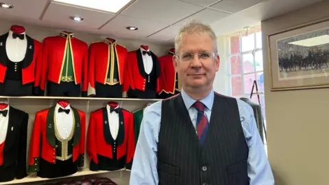 Lee Dawson stands in front of a row of military uniforms on mannequins on shelves behind him. The jackets are mostly red with gold trims and some with black or green cloth underneath.