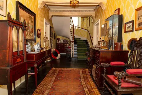 Steven Gourlay An old fashioned-looking interior of a building, featuring a winding staircase, chairs, sideboards, and an old rug on the floor.