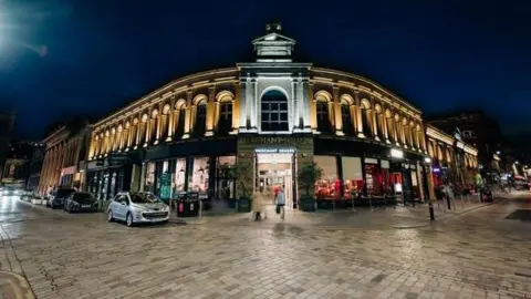 A large building, lit up at night, with people walking past it 