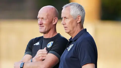 PA Media Paul Bastock wears a tight-fitting black sports T-shirt, and Ian Culverhouse, on the right, wears a navy blue polo shirt. They are outside, and are looking side-on to the left of the frame.