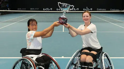 Getty Images Two female wheelchair tennis players lift a silver trophy between them as they smile for the camera with an empty tennis court behind them.