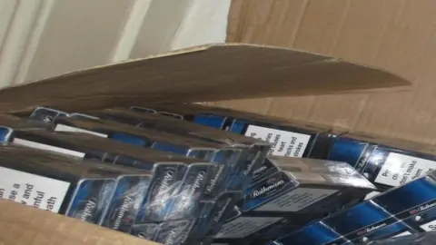 An open brown box filed with blue and white cigarette cartons.