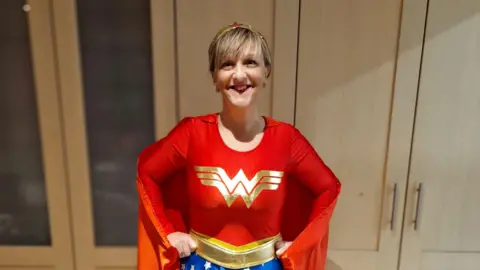 Family photo Marianne dressed up as Wonder Woman with a gold headband. She has her hands on her hips wearing a red cape and top with a golden W emblazoned on the front, with a gold belt and Wonder Woman's blue shorts with white stars just out of view. She is standing in front of some wooden cabinet doors.