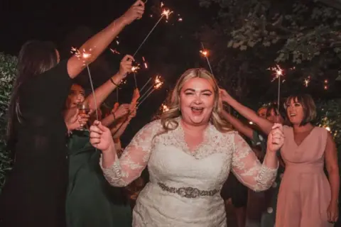 Neil Jones Photography A pistillate   successful  a achromatic  wedding formal  laughing astatine  the camera. She is holding a sparkler and determination   are guests down  her successful  dresses besides  holding sparklers 