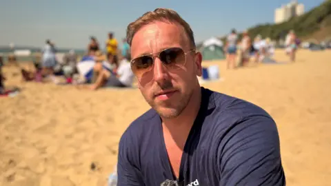 Liam Brownen, a man in a blue t-shire and sunglasses sat on the beach looking to the camera