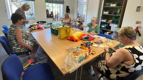 Five women sit around a long table which has bags and knitted items such as worm toys and blankets scattered across it. They are in a room with white walls. The women all wear sleeveless or short -sleeved floral dresses and sit on blue chairs. A store cupboard with items in green boxes can be seen. The sun shines outside the room's two windows.