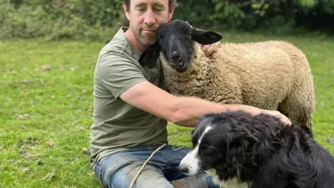 A man, sheep and a collie dog are huddled on a field. The man is hugging the sheep and is resting a hand on the dogs back.