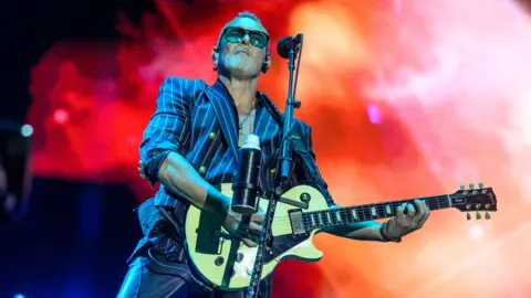 Getty Images Vivian Campbell, do Def Leppard, executa no palco. Ele está segurando um violão e usando um blazer despojado da Marinha com botões de ouro e óculos de sol. As luzes atrás dele estão vermelhas.