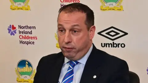 Pacemaker Roy McGivern speaks at a press conference. He has short black hair and is wearing a dark suit, light blue shirt and red, white and blue tie. He is sitting in front of a billboard with the Linfield FC crest, the Umbro logo and the NI Children's Hospice logo.
