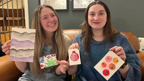 Poppy Barrett (left) and Hannah Broughton (right) are sitting on a brown leather sofa, each holding up small pieces of colourful artwork. The pieces include abstract purple waves, bright green leaves with a pink flower, a red pomegranate, and several circular fruit designs, possibly tomatoes of different colours. Behind them is a dark green wall with framed art.