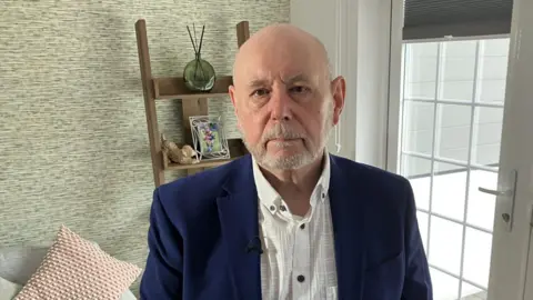 John Miller looks sombre and is wearing a navy suit jacket over a white shirt. He is stood in his lounge with a sofa and patterned cushions behind him.