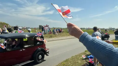 BBC A hand holding a Guernsey flag waving in the air as a WW2-era car drives past, with people lining both of sides of the road celebrating Liberation Day.