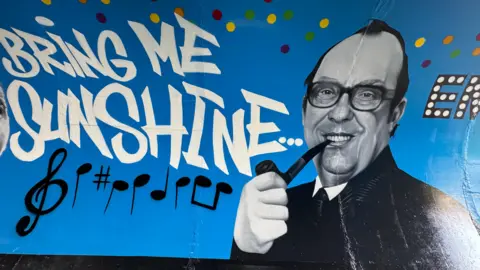 Hertfordshire County Council A painting of Eric Morecambe on a wall with lyrics to Bring Me Sunshine and musical notes written next to him