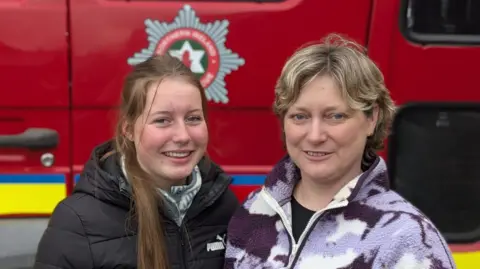 Gillian Thompson is wearing a purple and white camouflage fleece and her daughter Grace has long brown hair and is wearing a black coat and green and white fleece. Both are standing side-by-side beside a fire engine. 