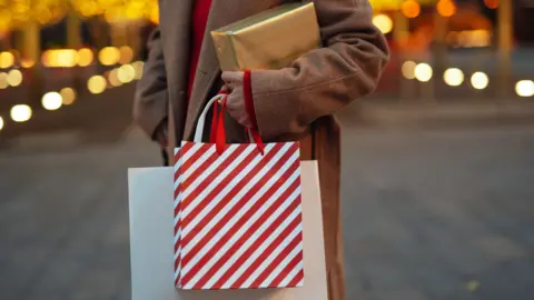 Getty Images A woman wearing a brown overcoat and red sweater stands in a street decorated for Christmas, holding a gold-wrapped parcel and two shopping bags.