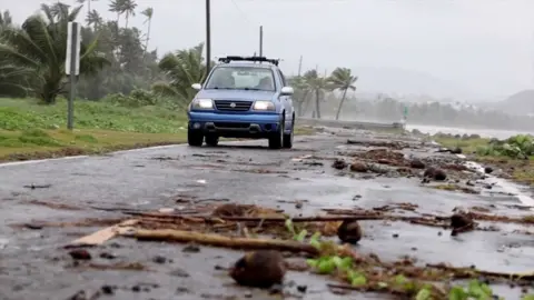 A bluish  car   drives on  a roadworthy  littered with branches adjacent  to the sea