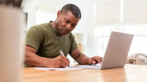 Getty Images A middle-aged veteran works at a laptop while writing on a piece of paper at a table. He wears a green t-shirt and a dog tag. 