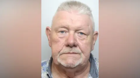 A police mugshot of a man in his 70s with a white beard and a blue shirt.