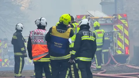 Firefighters, a safety officer and hazmat advisor stand in a group wearing high-vis firefighting attire and hard hats. The air around them is smoke-filled and multiple fire engines can be seen in the background.