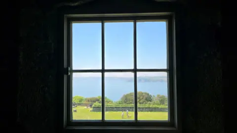 BBC Weather Watchers/ Silverloon A window in a dark room looks out onto a garden and the sea beyond it. 