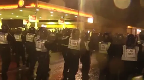 Hampshire and Isle of Wight Constabulary A large group of riot police can be seen making their way down a street under darkness. They are wearing helmets and high-visibility vest. They are just passing a Shell garage which has its forecourt illuminated in light. 