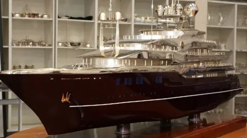 Elaborate model of a boat on a wooden stand. The bottom of the boat is carved from wood and the top, including the deck, radar and cabins are silver-coloured. 